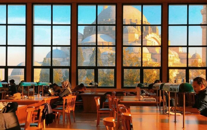 Interni accoglienti di una biblioteca con grandi finestre che mostrano una grandiosa architettura, e persone che leggono e studiano ai tavoli di legno. Interni accoglienti di una biblioteca con grandi finestre che mostrano una grandiosa architettura, e persone che leggono e studiano ai tavoli di legno.