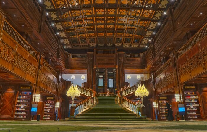 Interno grandioso di una biblioteca con una grande scalinata, pareti ornate in legno e scaffali pieni di libri in un'ambientazione magica. Interno grandioso di una biblioteca con una grande scalinata, pareti ornate in legno e scaffali pieni di libri in un'ambientazione magica.