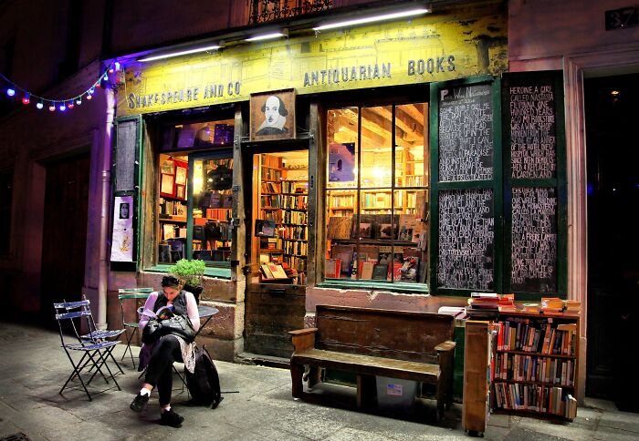 Cozy and grandiose bookstore at night with warm lighting, filled with books and a person reading outside. Cozy and grandiose bookstore at night with warm lighting, filled with books and a person reading outside.