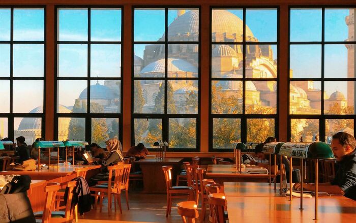 Cozy library interior with large windows showing grandiose architecture, and people reading and studying at wooden tables. Cozy library interior with large windows showing grandiose architecture, and people reading and studying at wooden tables.